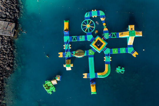 Aerial view of an inflatable water park in Gran Canaria with slides, balance beams, circular spinner, and central pyramid. People navigate and dive near a rocky breakwater at midday.