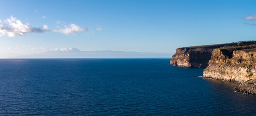 Rugged ochre cliffs of Gran Canaria meet the Atlantic, calm seas to horizon. Tenerife with Mount Teide appears left under a cloud band in late afternoon light.