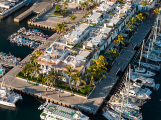 Aerial view of Puerto de Mogan on Gran Canaria shows whitewashed green trimmed houses, palm promenades, canals with sailboats, tidy walkways, and waterfront cafes in warm light. © Aerial Film Studio