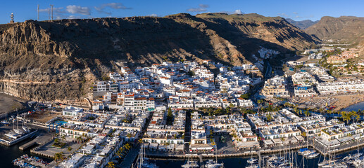 Aerial view of Puerto de Mogan, Gran Canaria, with white buildings, terracotta roofs, steep brown cliffs, palm promenades, moored sailboats, canals, sandy beach, and calm bay. © Aerial Film Studio