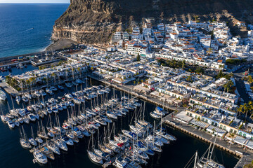 Aerial view of Puerto de Mogan, Gran Canaria shows a marina packed with sailboats, whitewashed buildings, palm lined streets, canals, quays, and steep volcanic cliffs in late afternoon. © Aerial Film Studio