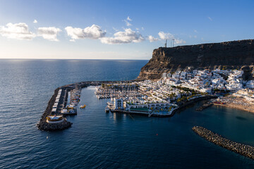 Aerial view of Puerto de Mogan, Gran Canaria, with marina, curved breakwaters, sailboats, sandy beach, white buildings with green trim, and antenna masts at late afternoon. © Aerial Film Studio