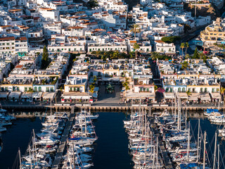 Aerial view of Puerto de Mogan, Gran Canaria, showing whitewashed buildings, green trimmed windows, sailboats in calm dark water, palms, cafes, and late afternoon light. © Aerial Film Studio