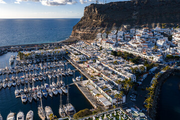 Aerial scene of Puerto de Mogan, Gran Canaria, with sailboats in a dense marina, whitewashed blocks, palm promenades, canals, piers, and a rugged cliff in late afternoon light. © Aerial Film Studio