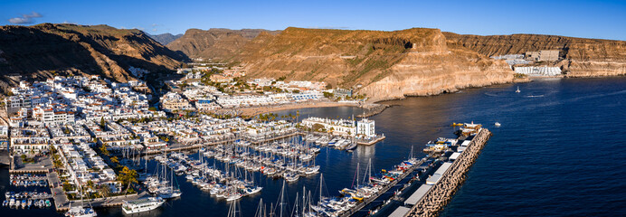 Aerial view shows Puerto de Mogan, Gran Canaria, with whitewashed flat roofs, marina of sailboats and yachts, breakwater, golden cliffs, beach, and canal like waterways. © Aerial Film Studio