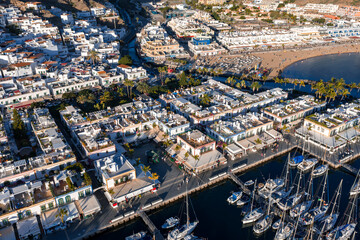 Aerial view of Puerto de Mogan on Gran Canaria shows marina sailboats, canal waterways, palm promenade, and busy crescent beach, set against terraced hills in warm light. © Aerial Film Studio