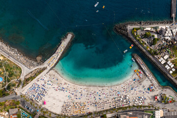 Aerial top down view of Amadores Beach, Gran Canaria, Spain. Curved seawalls enclose a lagoon with boats and pedalos, packed loungers, palm streets, and resort terraces. © Aerial Film Studio