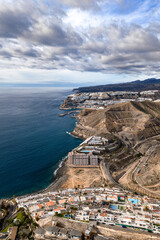 Terraced whitewashed resorts line arid cliffs above deep cobalt water near Puerto Rico and Amadores, with marinas, breakwaters, pools, and roads under soft afternoon light. © Aerial Film Studio