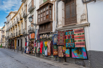 Naklejka premium A charming view of a traditional shop in Granada, Spain, displaying colorful Moroccan-style rugs, tapestries, and clothing against a backdrop of historic Andalusian architecture.