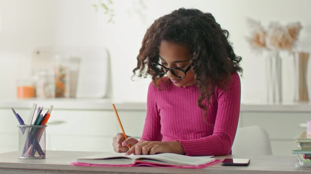 A girl writes in her notebook at the kitchen table while studying for school. She uses a pencil and appears focused on her schoolwork during the day.