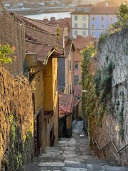 A cozy street view in Porto, Portugal during sunset. Old houses with red tile roofs stand between stone walls, leading to colorful buildings near the river. 