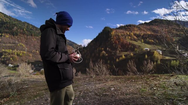Man adjusting drone controller and settings while checking smartphone telemetry, focused expression and steady hands. Coldweather jacket and beanie, remote firmware and battery checks, detailed prep