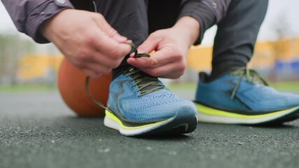 athlete tying blue running shoe, closeup of hands knotting laces with basketball tucked under knee on outdoor asphalt court, morning light, casual jacket and leggings, preworkout warmup, focused