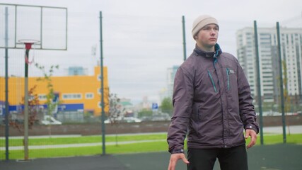 Man dribbling basketball on court, overcast urban park with hoop and chainlink fence, wearing beanie and puffer jacket, solitary athlete practicing focused moves, residential towers in background,
