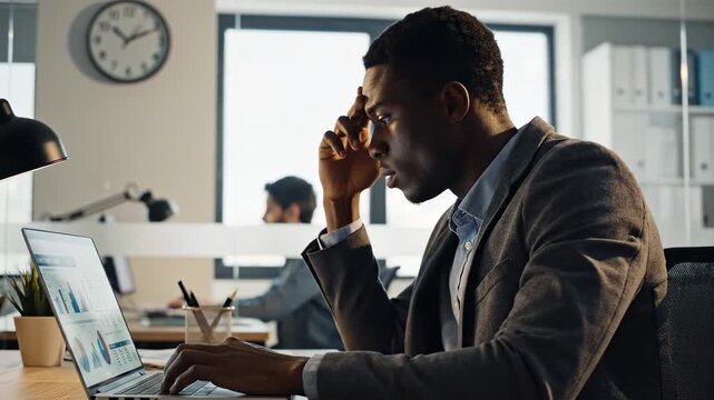 An African American businessman sits at his modern office desk, looking frustrated and holding his head in a moment of stress. His laptop displays various charts and graphs, indicating a demanding