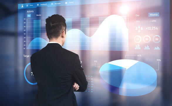 A businessman stands in front of a digital screen, analyzing interactive graphs and charts. The modern office setting highlights the importance of data analysis in decision-making. Impute - Powered by Adobe
