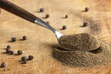 Ground black pepper on a stainless steel spoon with whole peppercorns scattered on wooden surface.