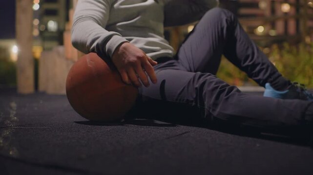 night court player resting with basketball, urban rooftop court lit by warm city lights, male athlete wearing hoodie and pants sits on asphalt, hand gripping ball, sneaker sole visible, contemplative