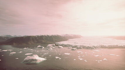 In a tranquil Arctic fjord, icebergs drift peacefully while the sun sets. The landscape is adorned with distant mountains, casting soft reflections on the shimmering water. © icetray