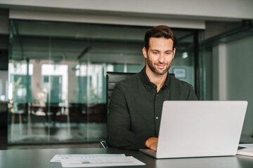 Young professional it specialist European business man working on laptop pc sitting at desk in modern office. Middle-aged latino businessman using computer technology app for work online. Copy space