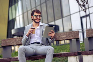 Young businessman sitting on a bench outside a modern office building, enjoying coffee while using a tablet. Ideal for urban lifestyle, business, technology, and professional concepts.