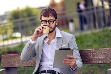 Young businessman sitting on a bench outside a modern office building, enjoying coffee while using a tablet. Ideal for urban lifestyle, business, technology, and professional concepts.