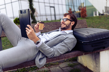 Young businessman lying on a bench outdoors, drinking coffee and reviewing a paper report.