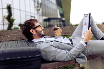 Young businessman lying on a bench outdoors, drinking coffee and reviewing a paper report.