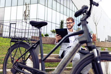 Smiling young businessman holding a tablet while standing next to his bicycle in a city environment.