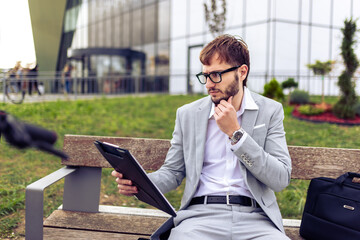 Young businessman sitting on a bench outdoors, carefully reviewing a paper report.