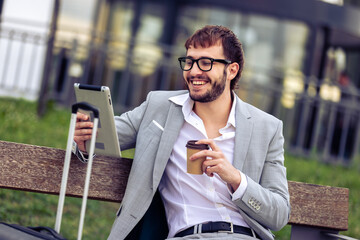 Young businessman sitting on a bench in front of a modern office building, drinking coffee and using a tablet.