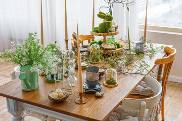 Stylish Easter table decor with candles, greenery, bunny figurines and natural elements in bright spring room interior. Cozy festive dining setup with wooden table, soft daylight and seasonal details