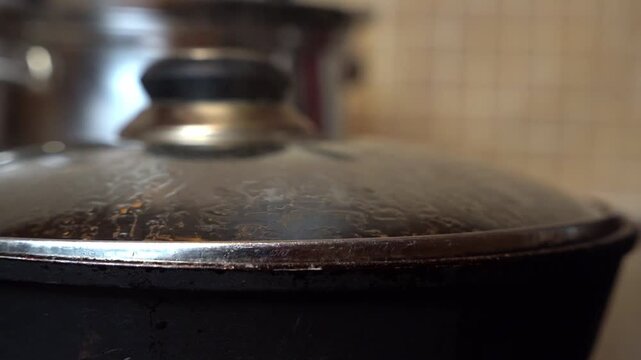 A close-up of a black cooking pot with a metallic lid. The background features a blurred silver pot and a tiled kitchen wall.