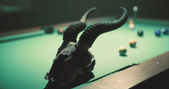 A skull with prominent horns is positioned on a pool table. Surrounding it are colorful billiard balls and a soft, atmospheric light casts shadows, enhancing the mysterious vibe of the setting.