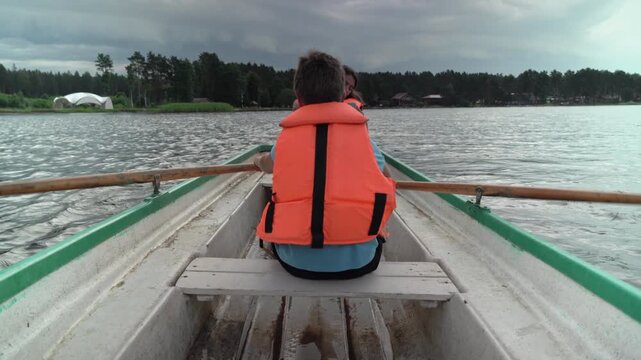 A young boy with brown hair wearing an orange life jacket rows a small boat on a calm lake. Dark clouds loom in the background, hinting at an approaching storm.