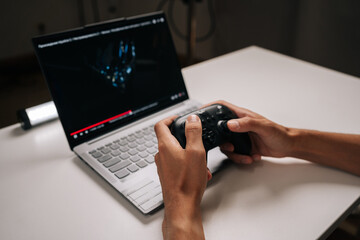 Person playing video game on laptop using wireless controller, hands gripping gamepad, immersed in digital entertainment and online gaming, digital leisure activity. © dikushin