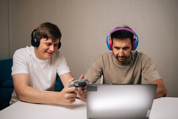 Happy excited young men gathered in living room, laughing and focused on laptop and controllers while gaming together, showcasing friendship, teamwork and casual digital leisure.