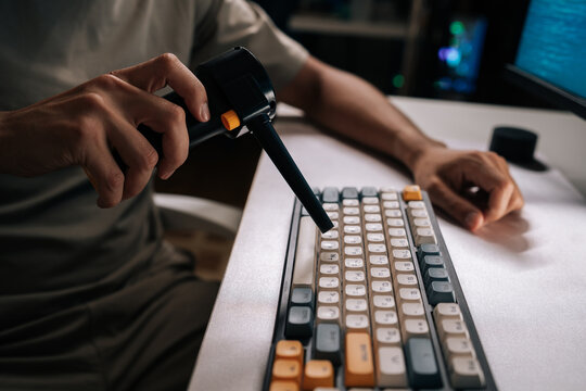 Person cleaning computer mechanical keyboard on white desk, using electric air duster to remove dust and debris from keys as part of regular maintenance and tech hygiene.