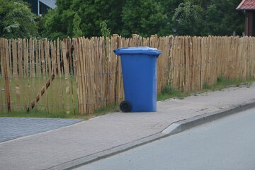 Naklejka premium A blue, closed waste bin for paper waste is ready for collection by the wooden fence on the sidewalk