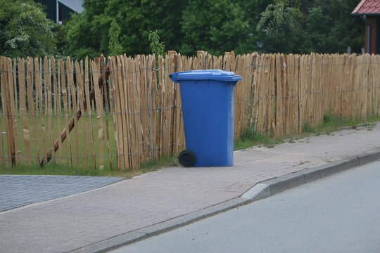 A blue, closed waste bin for paper waste is ready for collection by the wooden fence on the sidewalk