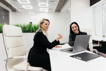 Two professional women engaged in a discussion at a modern office workspace with a laptop on a white table and greenery in the background during a collaborative meeting