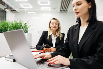 Two businesswomen in black suits working in a modern office with a laptop and smartphone, greenery in the background, showcasing a collaborative work environment