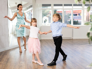 Fototapeta premium Positive preteen girl and boy practicing samba steps in latin dance studio while female teacher observing and guiding couple..