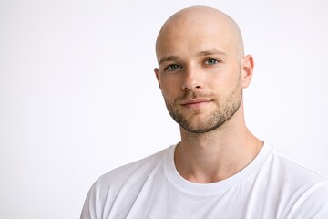 Obraz premium Close-up portrait of a handsome bald man with green eyes and light stubble wearing a white t-shirt against a bright white background.