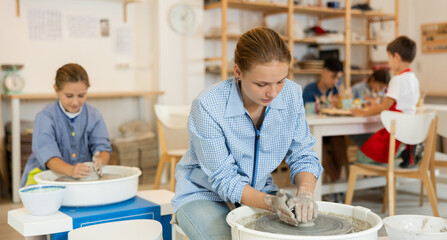 Obraz premium Concentrated teenage girl working at potter's wheel in taller workroom with other circle members
