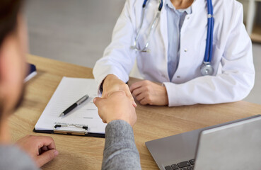 Handshake of doctor and patient during hospital visit. Female physician with stethoscope and man shake hands over clipboard with medical notes on desk, building trust and deal agreement on meeting