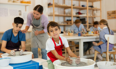 Boy in an apron is sculpting a mug on a potter's wheel. Child at a ceramic workshop with a teacher...