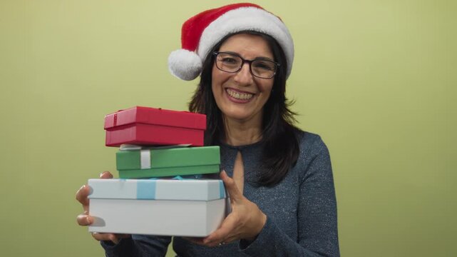 Woman wearing santa hat and glasses holding three colorful gift boxes in studio with pale green background; joy.