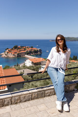 Young woman in casual clothes sitting on the railing of the observation deck overlooking the sea and the island in the background, summer vacation, travel and tourism concept, space for text