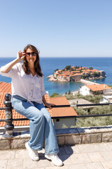 Young woman in casual clothes sitting on the railing of the observation deck overlooking the sea and the island in the background, summer vacation, travel and tourism concept, space for text